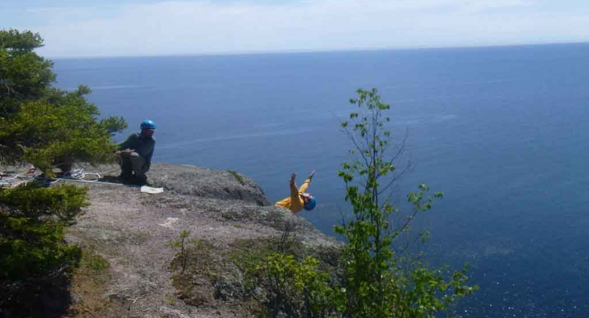 a person who is attached to ropes while rock climbing leans back and throws their hands back over a vast blue body of water